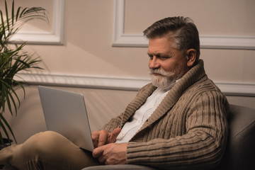 happy senior man using laptop while sitting in armchair