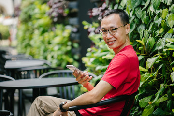 A middle-aged Asian man is sitting on a row of tables on a quiet afternoon. with phone in city. He is smiling nicely as he patiently waits for his friend.