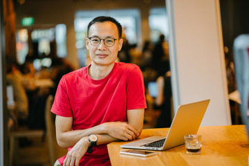  A portrait of a n attractive Asian Chinese dressed comfortably in a loose shirt and pants. He is sitting inside a cafe while working on his laptop with his smartphone in front of him.