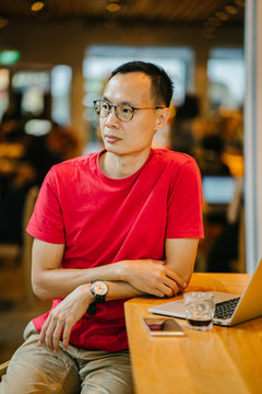 An image of a serious looking middle age Asian guy is sitting insiode a cafe while working on his laptop. He is looking so fierce and concentrated while looking away from the camera.