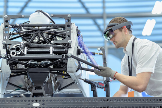 Engineer using augmented reality headset to 'see' parts position on car in assembly composite image showing CAD drawing of part in robotics research facility