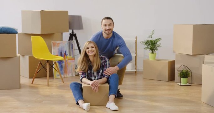 Young happy good looking couple sitting on the floor and posing in front of the camera and smiling among many boxes in their new home while moving in it. Portrait shot. Inside