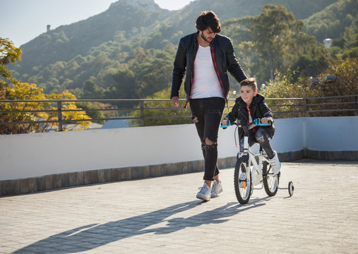 Young boy riding bike with stabilizers, father walking beside him