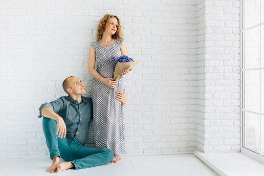 Man sitting barefoot on the floor hugging his pretty pregnant wife in long dress holding bouquet.