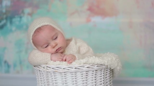 Little cute baby boy, dressed in handmade knitted white teddy bear overall, sleeping peaefully with little knitted toy in basket