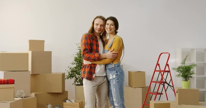 Young happy just married man and woman standing in the middle of the cozy living room surrounded by unpacked boxes and posing to the camera while hugging. Indoor