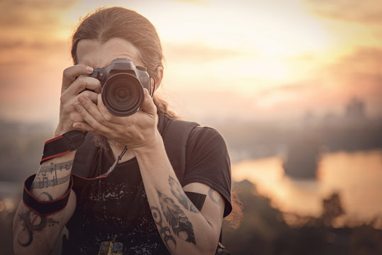 Young photographer with long hair and alternative style taking photographs with his dslr camera, capturing landscape and sunset in a park