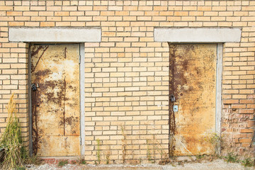 The texture of the old brick wall with concrete seams, green grass near the ground and two old rusty doors on a clear sunny day.