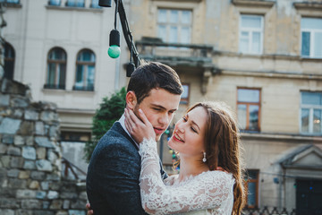 Fototapeta premium Wedding couple hugs in the old city. Rustic bride with hair down holds face of a groom in grey suit and bow tie. Love in ancient medieval town. White lace wedding dress. Vintage architecture details.