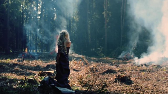 Dramatic young woman stands on the background of a burning forest and looks on bonfires. Stylish girl is standing motionless and looking at the smoke and ash