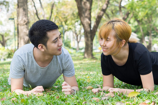 Asian sweet couple exercising together with smile on their faces on soft grass encircle with nature and warm light from the sun in the afternoon. Couple smile happily to each other. Outdoor exercise.