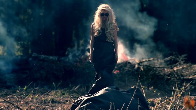A young woman comes to the fore plane and stands on the background of a bright fire and forest. Scenic view with smoke and ashes. Dramatic passage of freaky blond girl