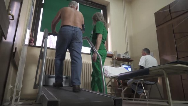 Test With Workload Of The Heart. Cardiologist Testing Heart Rate Of Man Who Walking On Treadmill, Doctor Watching Result On Computer Screen And Nurse Assisting Patient, Cardiology Ward In Hospital