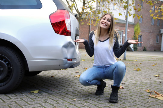 H&uuml;bsche blonde Frau sitzt an ihrem Auto mit Unfallschaden und lacht