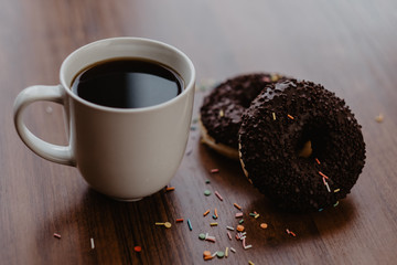 Black coffee in a white mug and two chocolate donuts on a wooden table by the window