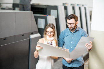 Young woman designer and print operator working together with paper print standing at the print...
