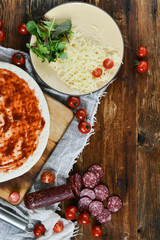 Ingredients for Pizza: basis dough, salami sausage, cherry tomatoes, grated cheese, tomato paste, herbs and arugula. Natural Wooden Background With Linen Napkin and Knife For Pizza