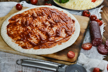 Ingredients for Pizza: basis dough, salami sausage, cherry tomatoes, grated cheese, tomato paste, herbs and arugula. Natural Wooden Background With Linen Napkin and Knife For Pizza