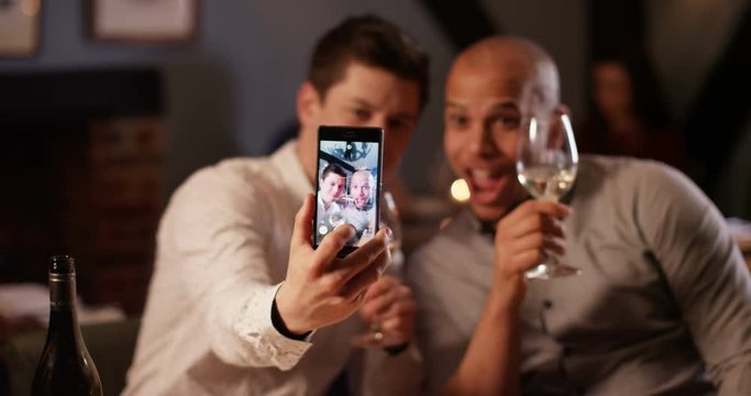 4K Happy male friends enjoying glass of wine on an evening out pose to take a selfie with smartphone