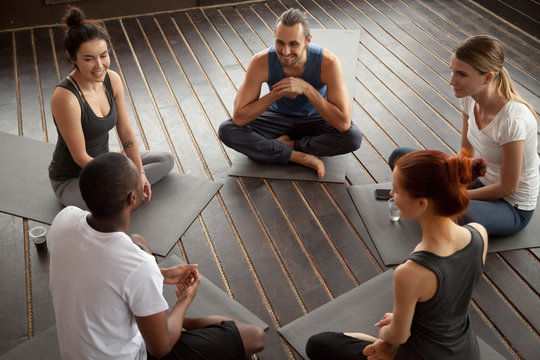 Diverse smiling people sitting on mats in circle relaxing talking before yoga training, happy multiracial friends group discuss healthy lifestyle motivation after pilates fitness class in gym studio