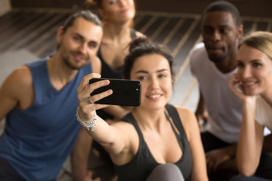 Taking selfie on smartphone concept, sporty woman holding cell shooting video or making diverse friends group self-portrait photo on modern smartphone camera at training break in gym, focus on phone