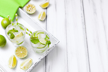 Caipirinha, mojito cocktail with lime, brown sugar, ice and mint leaves in beautiful glasses, cut green citrus on white wooden background. Summer alcohol drink. Close up photography. Selective focus