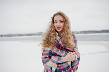 Curly blonde girl in checkered plaid against frozen lake at winter day.