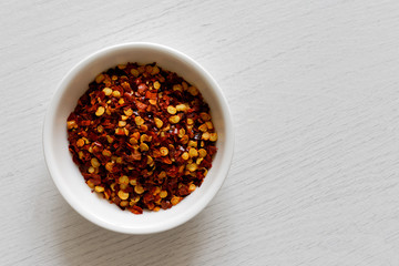 Coarsely ground chilli peppers in white ceramic bowl isolated on white wood background from above.