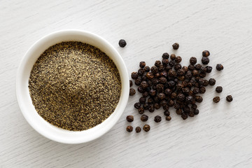 Finely ground black pepper in white ceramic bowl isolated on white wood background from above. Black peppercorns.