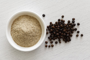Finely ground white pepper in white ceramic bowl next to black peppercorns isolated on white wood background from above.