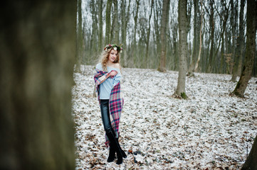 Curly cute blonde girl with wreath in checkered plaid at snowy forest in winter day.