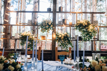Fototapeta na wymiar Table with blue tablecloth decorated with flowers and green branches, candles in tall vases next to table, light bulb garland on background