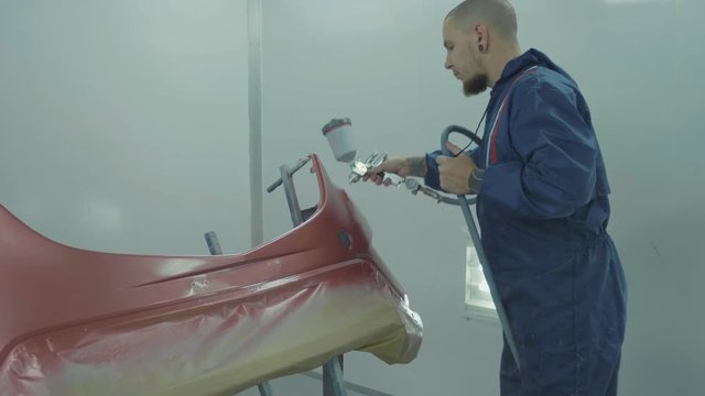 Auto painter spraying red paint on car front bumper in special booth
