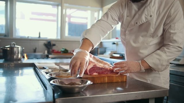 Butcher adding spices into large piece of fresh raw meat lying on a wooden board in a commercial kitchen, slow motion