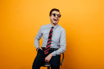 Happy cheerful smiling young businessman sitting on chair and wearing sunglasses over orange...