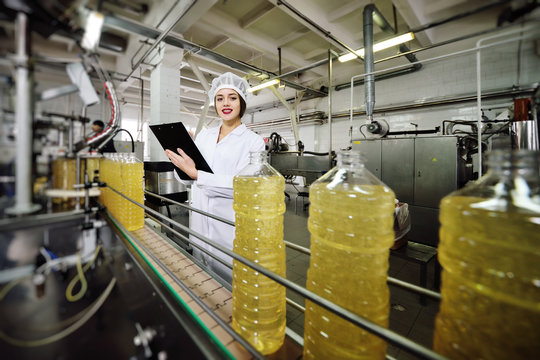 Beautiful Young Girl Is Making Notes On A Sheet Of Paper On The Background Of A Line On Food Production Of Sunflower Oil.