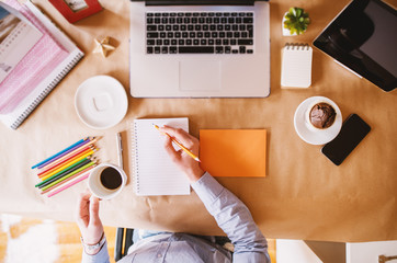 Top view of woman designer writing organization plans in notes while having a coffee break at the office desk.