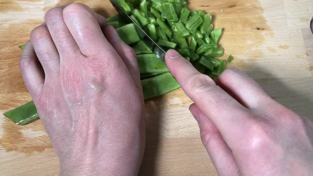 Close up of Caucasian female hands cutting slices of green Italian taccola, a kind of pea (Pisum sativum), uncooked on a wooden cutting board in a kitchen with a knife. Fast Version.