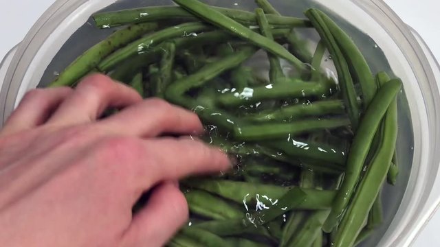 Raw green string beans on water in a plastic bowl on white background.