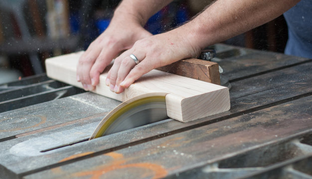 A close up photo of a man sawing through a piece of wood on a table saw with sawdust flying.