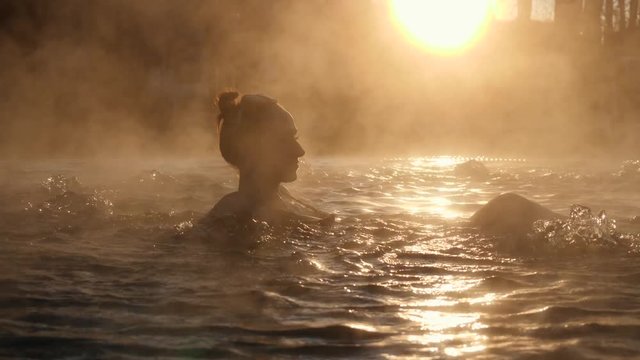 Geothermal spa. Woman relaxing in hot spring pool outdoors. Girl enjoying bathing in a blue water lagoon tourist attraction at sunset. Slow motion