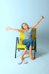 Cute Young Girl, Wearing blue Jeans And Striped T-Shirt, Sitting in Yellow Arm-Chair Over Blue Background and Holding Hands Up. 