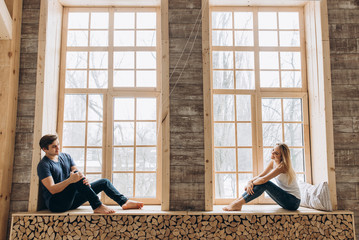 Happy guy and girl are sitting on the windowsill near the big window and look at each other lovingly