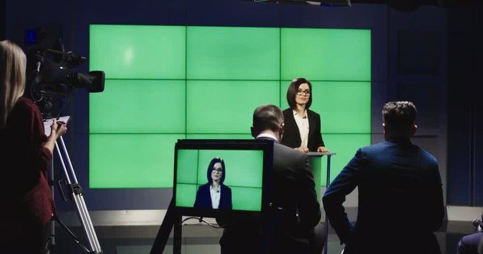 Young female politician in elegant suit standing on podium in studio against green chromakey screen and talking to press journalists.