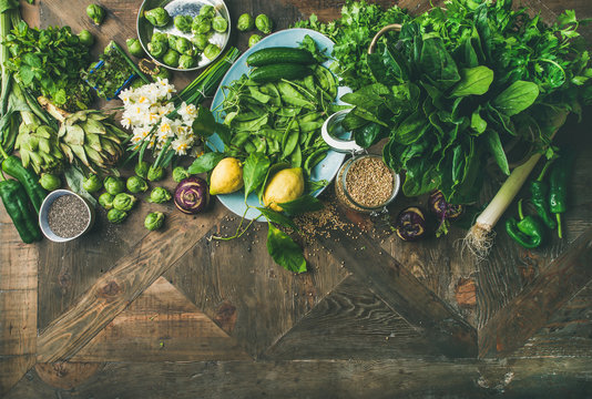 Spring healthy vegan food cooking ingredients. Flat-lay of vegetables, fruit, seeds, sprouts, flowers, greens over rustic wooden background, top view, copy space. Clean eating food concept