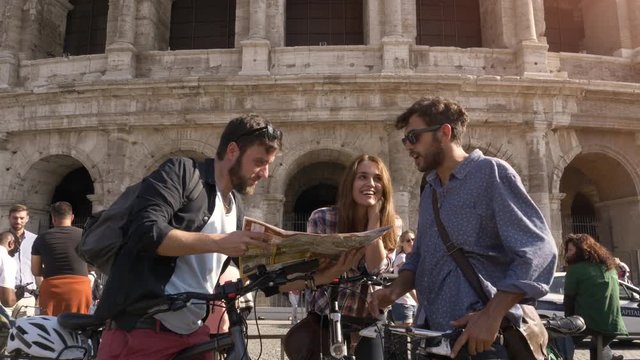 Three happy young friends tourists with bikes and backpacks at Colosseum in Rome reading map guide for directions on sunny day slow motion steadycam ground shot