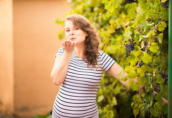 Beautiful young pregnant woman is standing in the summer near the grapes and sends an air kiss