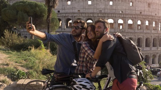 Three happy young friends tourists with bikes and backpacks at Colosseum in Rome taking selfies on hill at sunset with trees slow motion steadycam