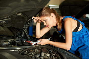 Brunette girl mechanics repairing or inspecting a car and holds a spanner in her hand. Girl is dressed in working clothes