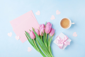 Coffee cup, spring tulip flowers, gift box and pink paper card on blue pastel table top view. Greeting for Womans or Mothers Day. Flat lay.